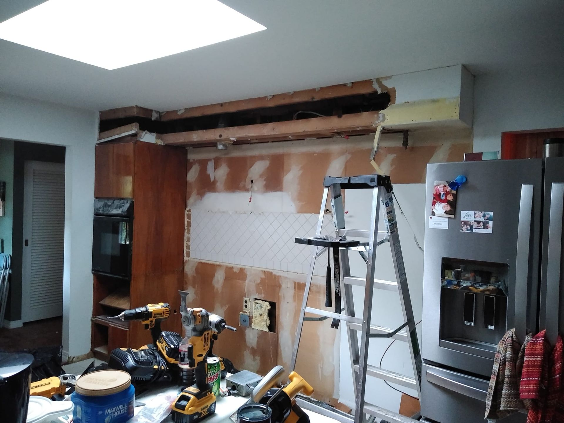 A kitchen renovation scene showing a partially opened wall with exposed studs, a step ladder, and tools on a counter.