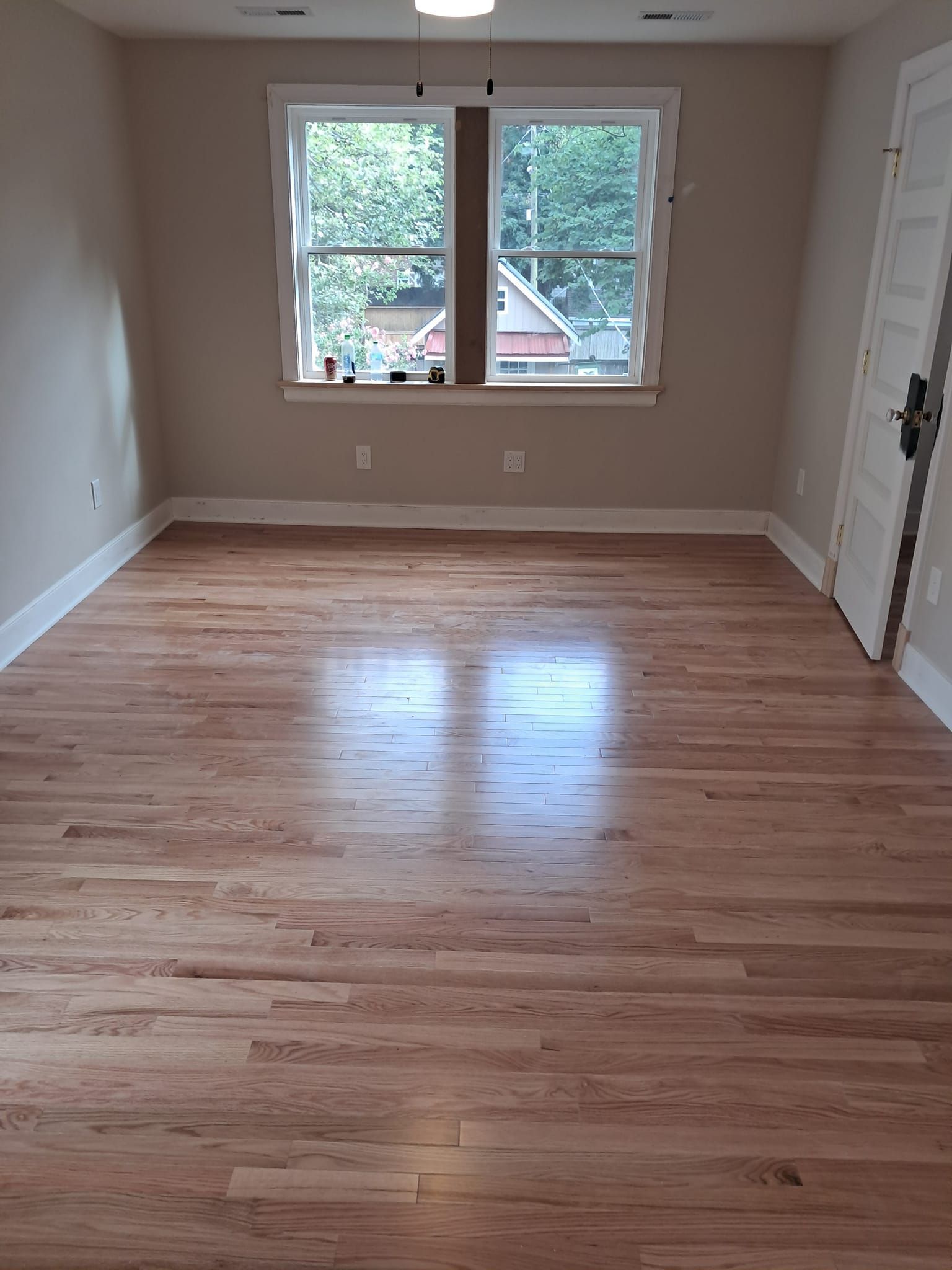 An empty room with polished light wood floors, beige walls, white trim, and a double window overlooking trees.