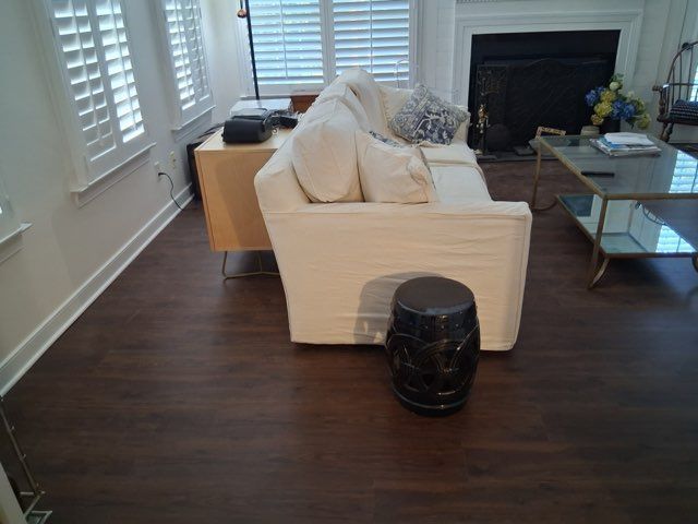 A living room features a beige sofa, dark wood flooring, a coffee table, and a small black stool in the foreground.