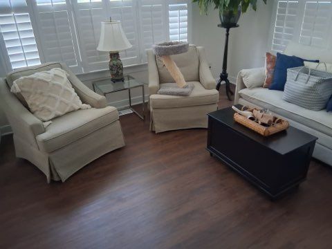 A bright living room with two beige armchairs, a white sofa, a glass side table, a black trunk coffee table, and a plant.