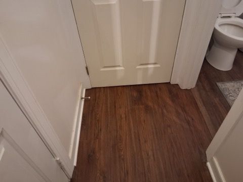 A hallway floor with dark wood-grain planks, showing a closed white door and a portion of a bathroom toilet.