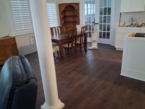 A living room with dark wood floors, a leather chair, a dining table with chairs, and white cabinets in the background.