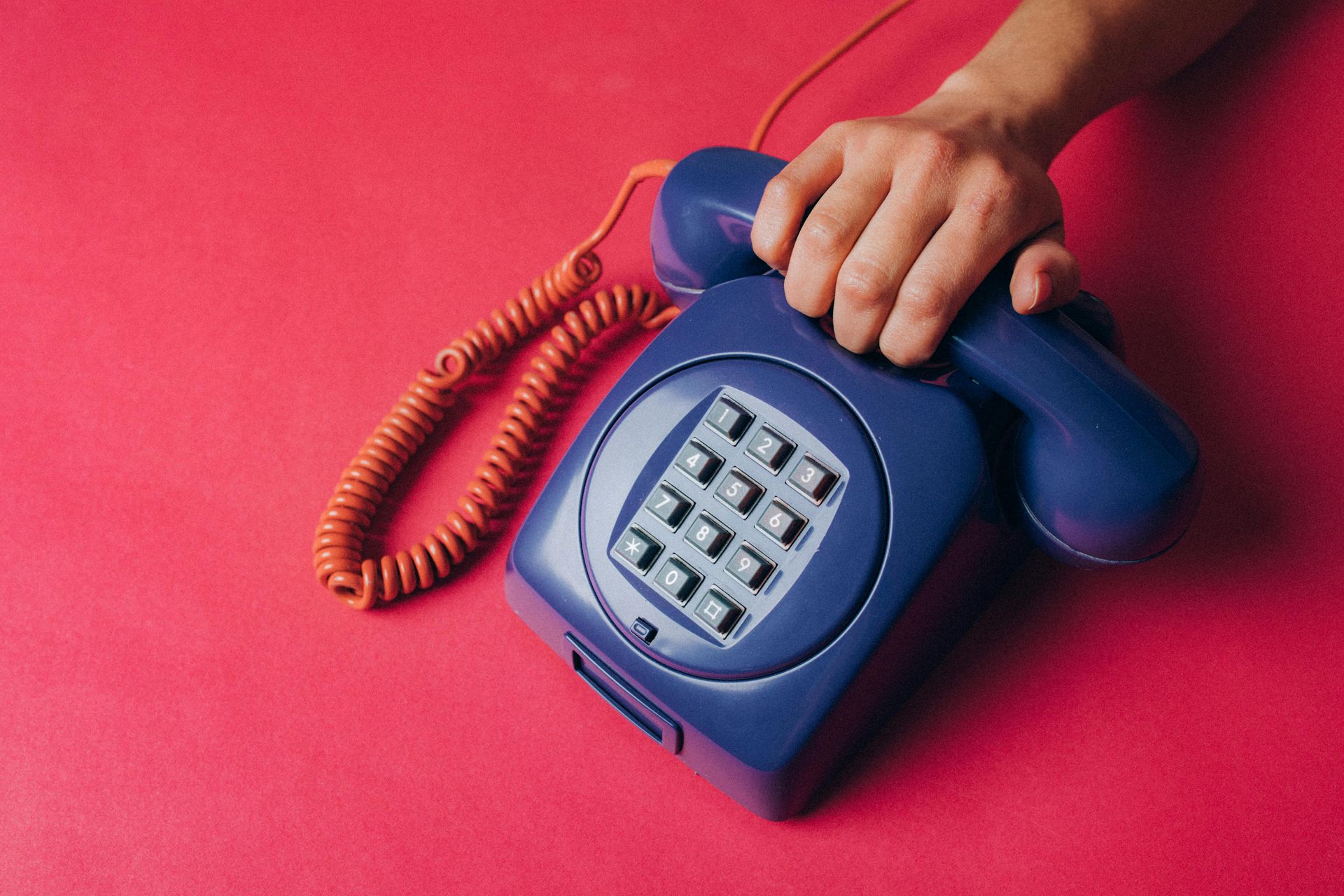 A hand holds the handset of a dark blue, push-button telephone against a vibrant red background.
