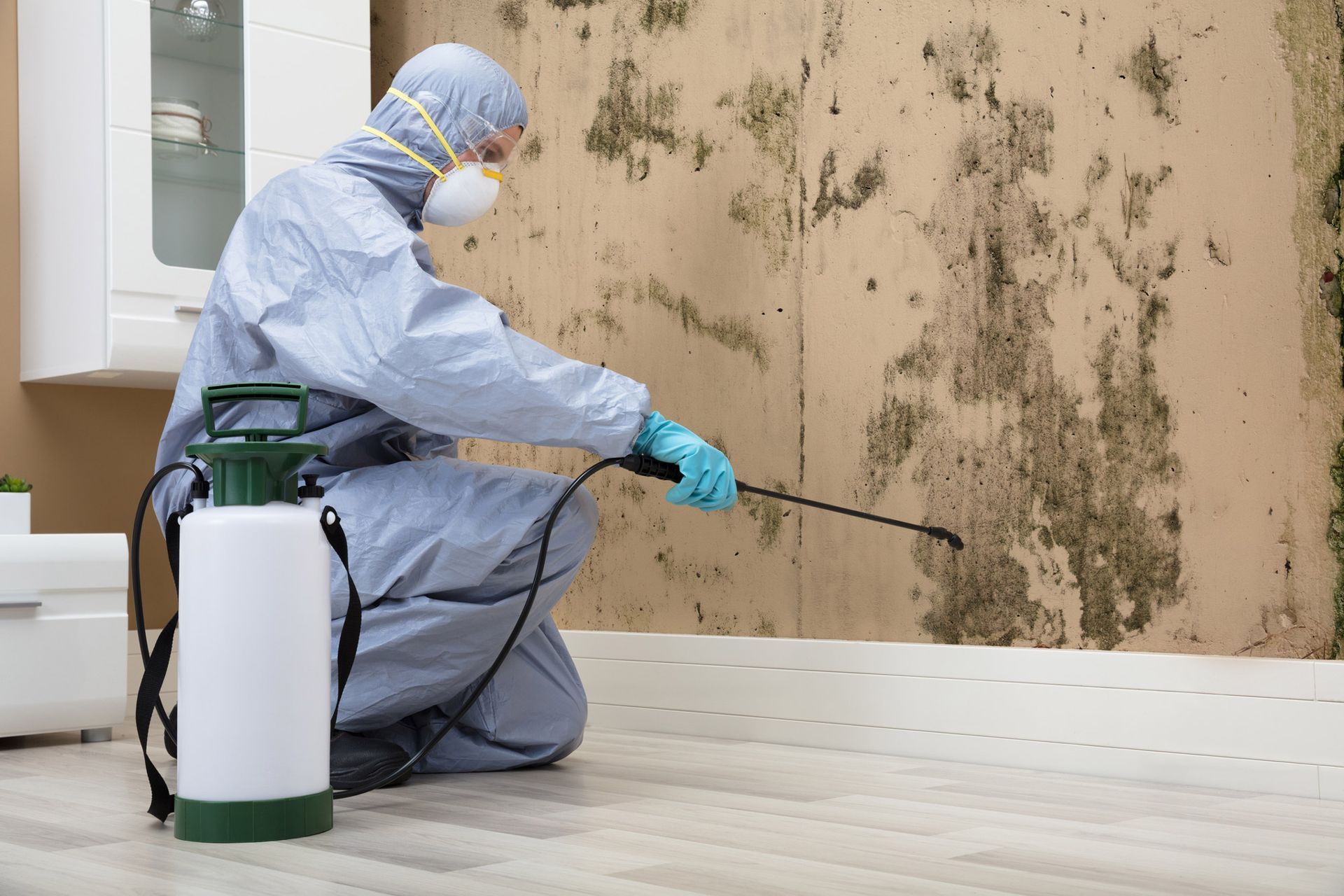 Pest control company worker in safety uniform sprays pesticide on wall inside a room.
