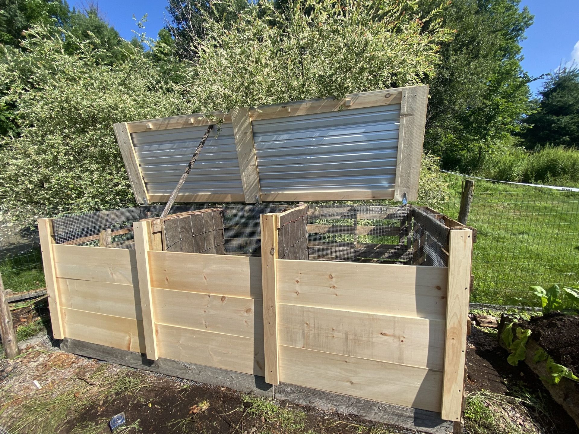 A wooden compost bin with the lid open is sitting in the grass.