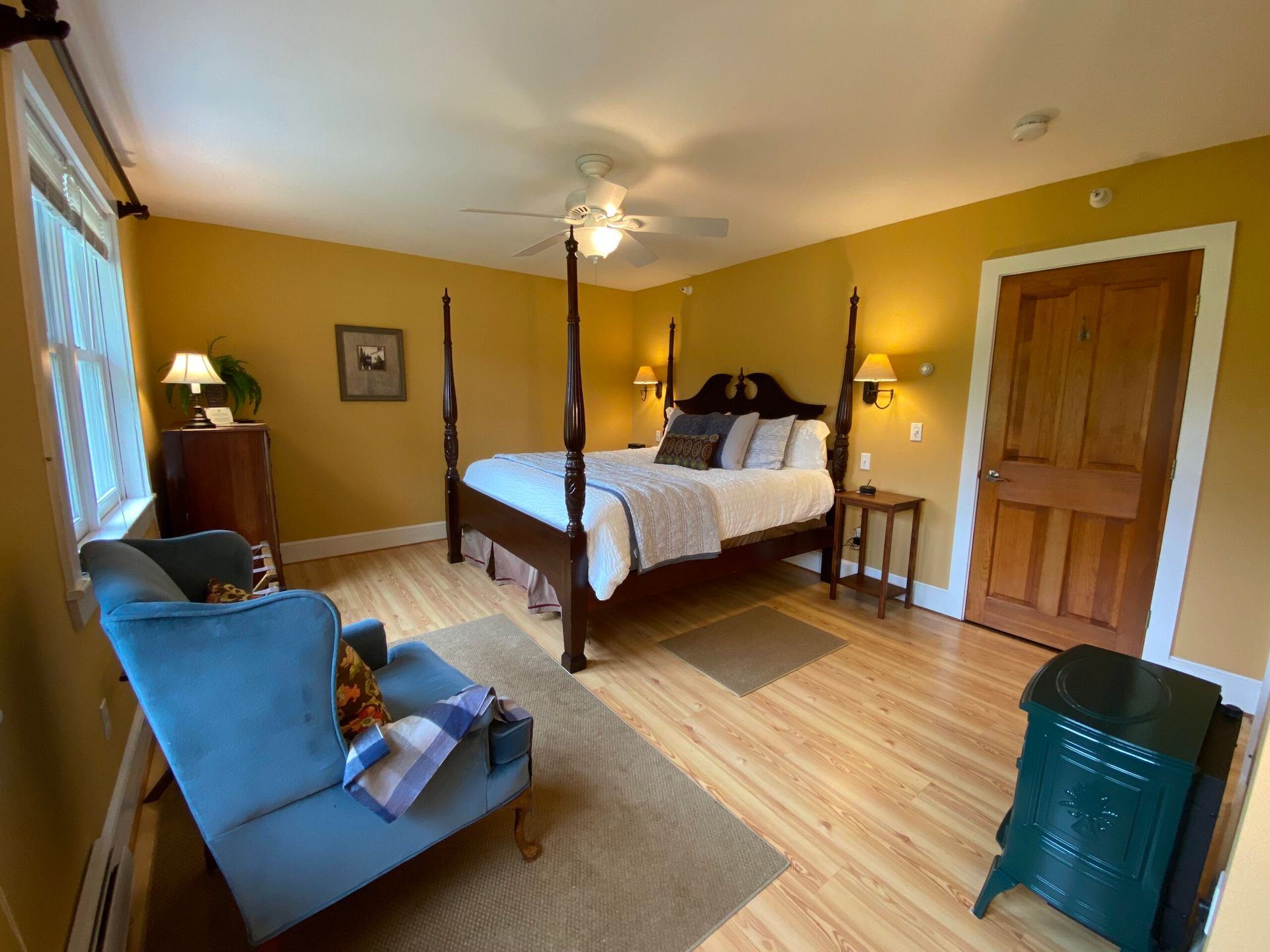 A bedroom with a four poster bed , chair , nightstand and ceiling fan.