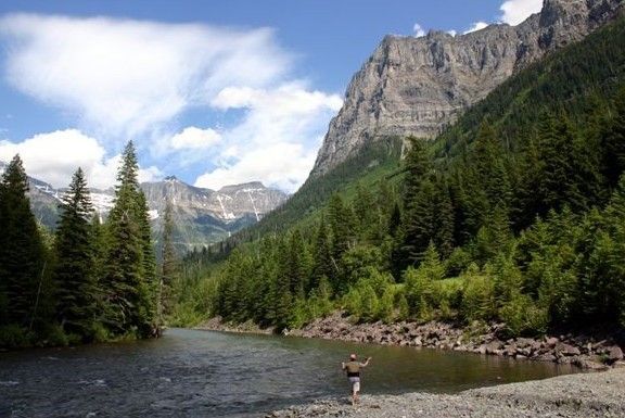 A man is fishing in a river with mountains in the background.