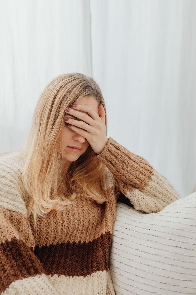 Woman seated on a couch, covering her face with one hand in a softly lit room.