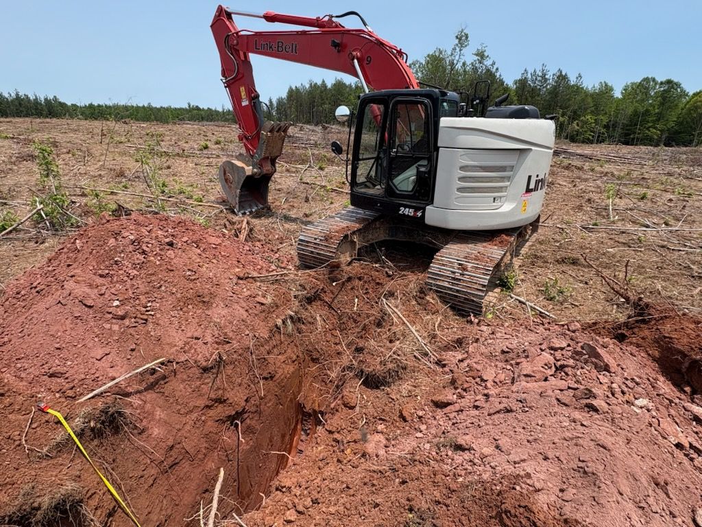 Excavator digging a trench in red soil on a cleared field, under a bright, sunny sky.