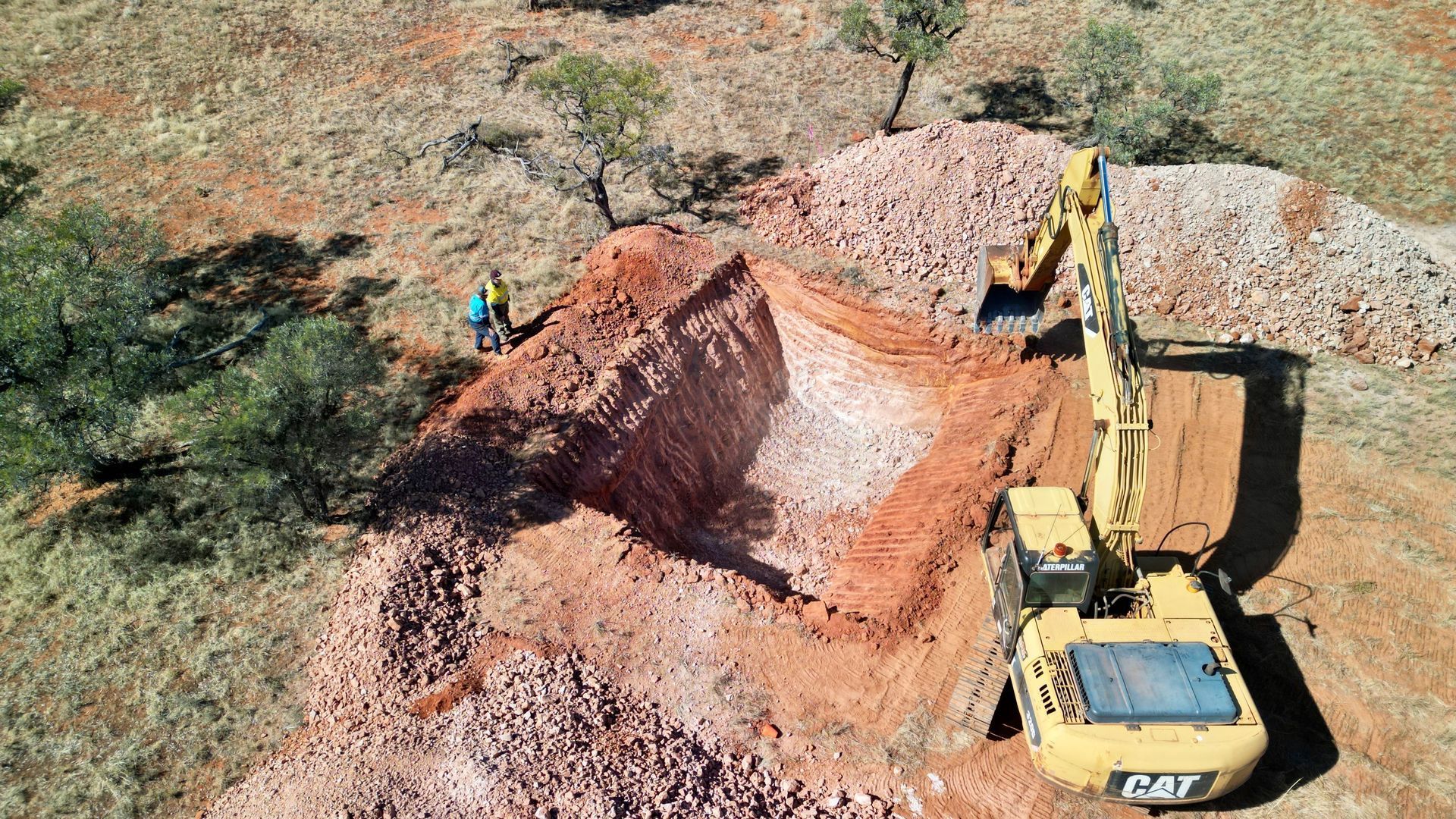 Excavator digging in a red dirt area; a person stands nearby. Piles of rocks surround the site.