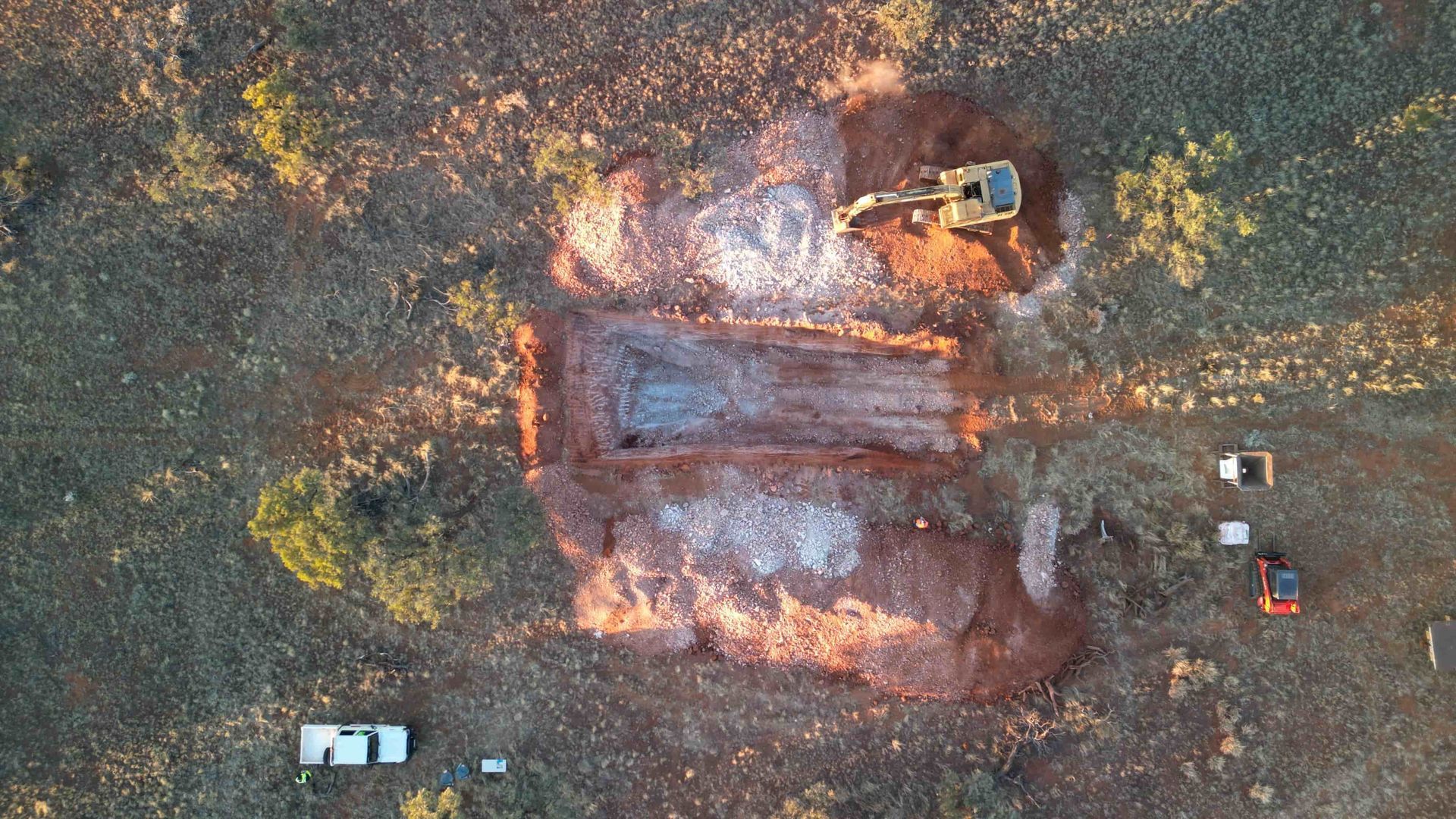 Aerial view of construction site with excavator digging a rectangular pit in a brown, arid landscape.