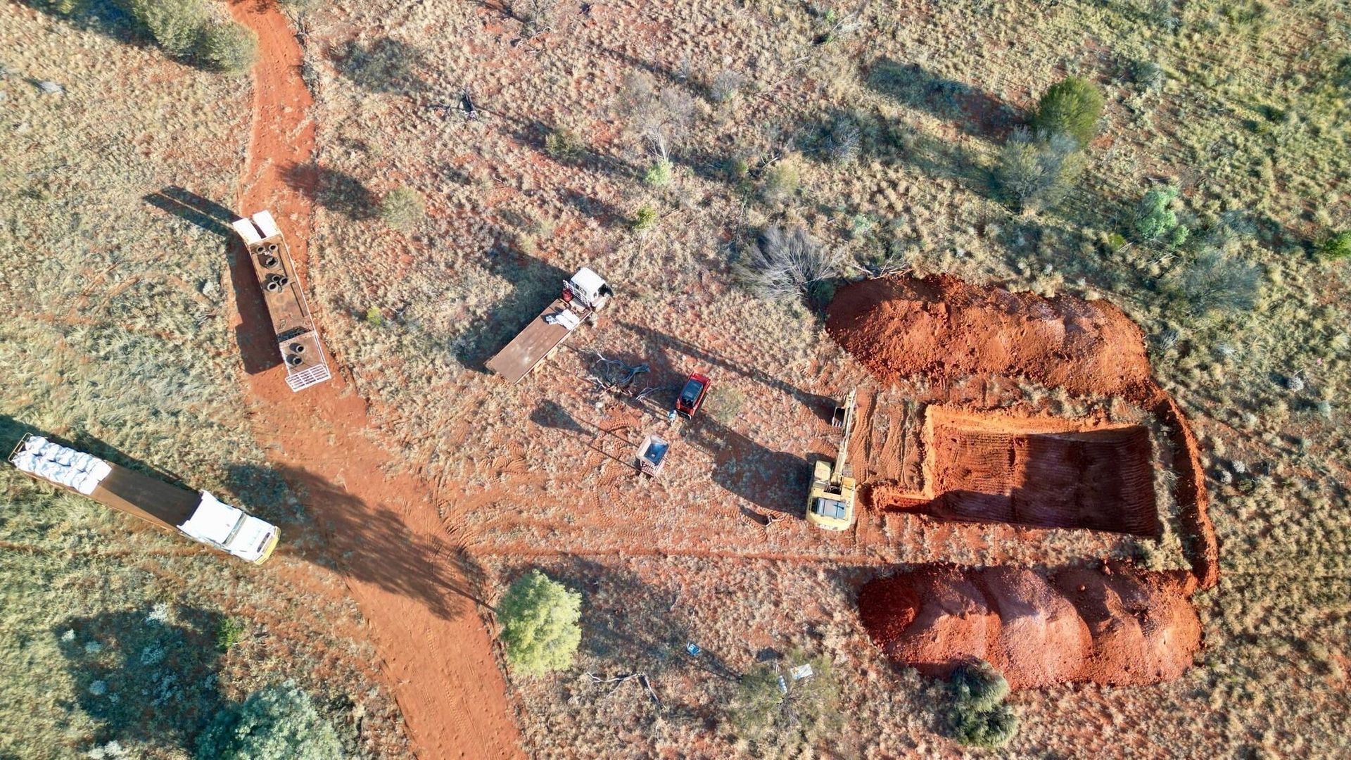 Aerial view of an excavation site in a reddish-brown desert landscape. Trucks, equipment, and workers are present.