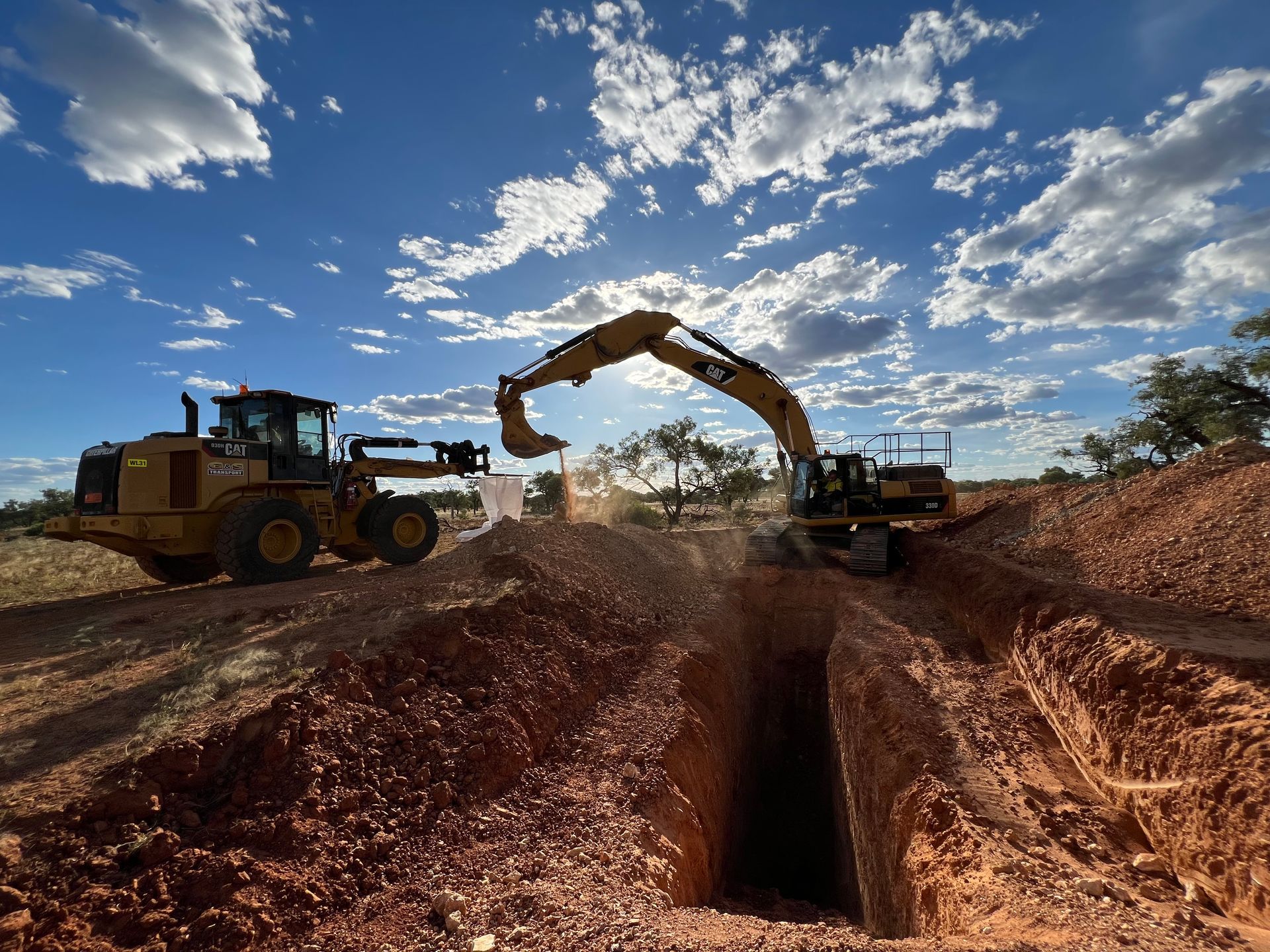Excavators digging a trench in reddish soil under a blue sky, with a front-end loader nearby.
