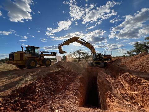 Excavators digging a trench in reddish soil under a blue sky, with a front-end loader nearby.