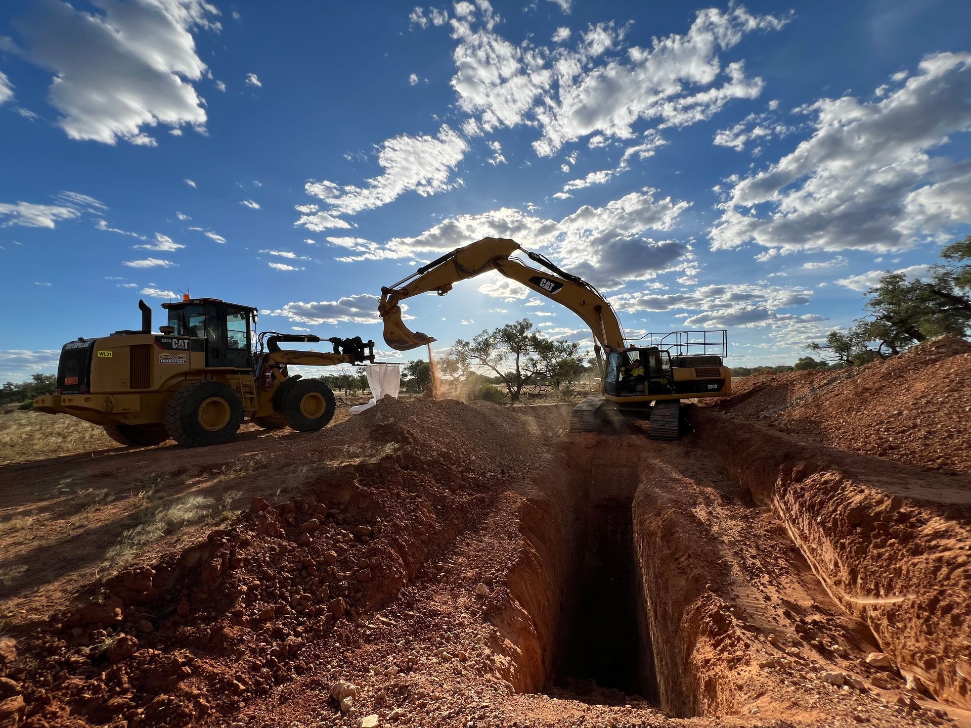 Excavators and loader digging a trench in reddish soil under a blue sky, sunlight filtering through.