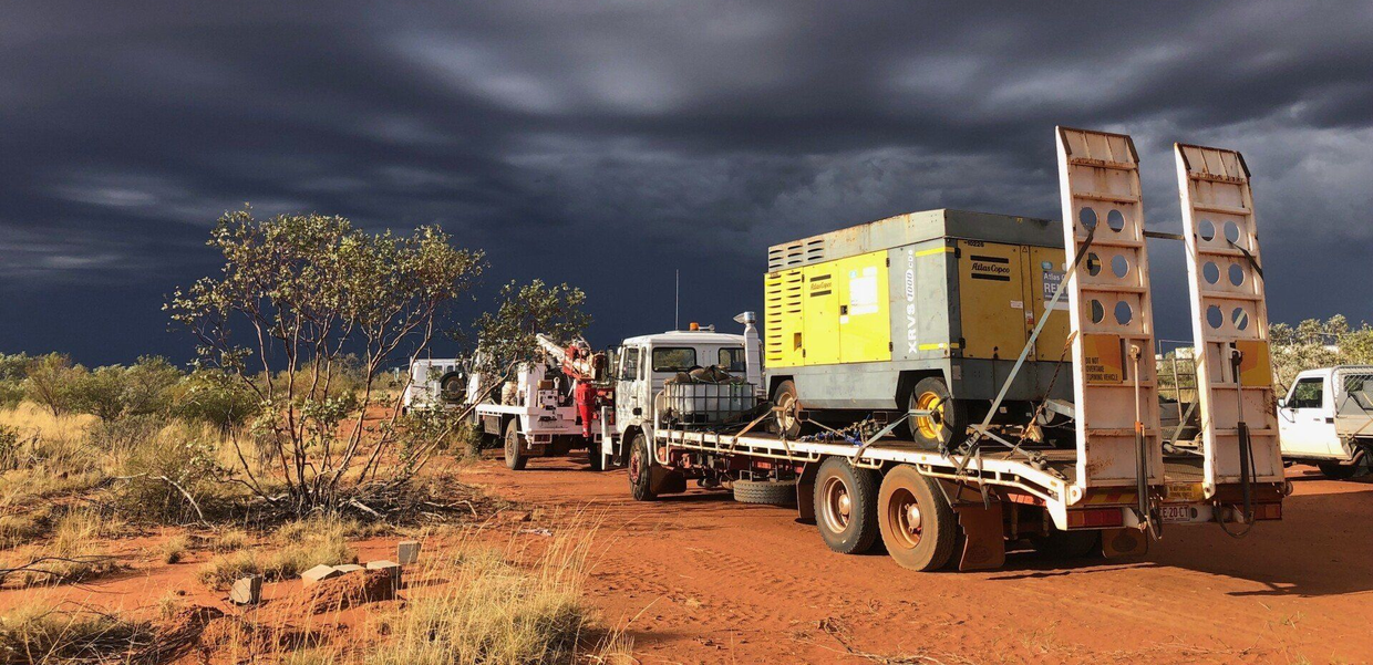 Trucks carrying industrial equipment on a red dirt road under a dark, stormy sky.