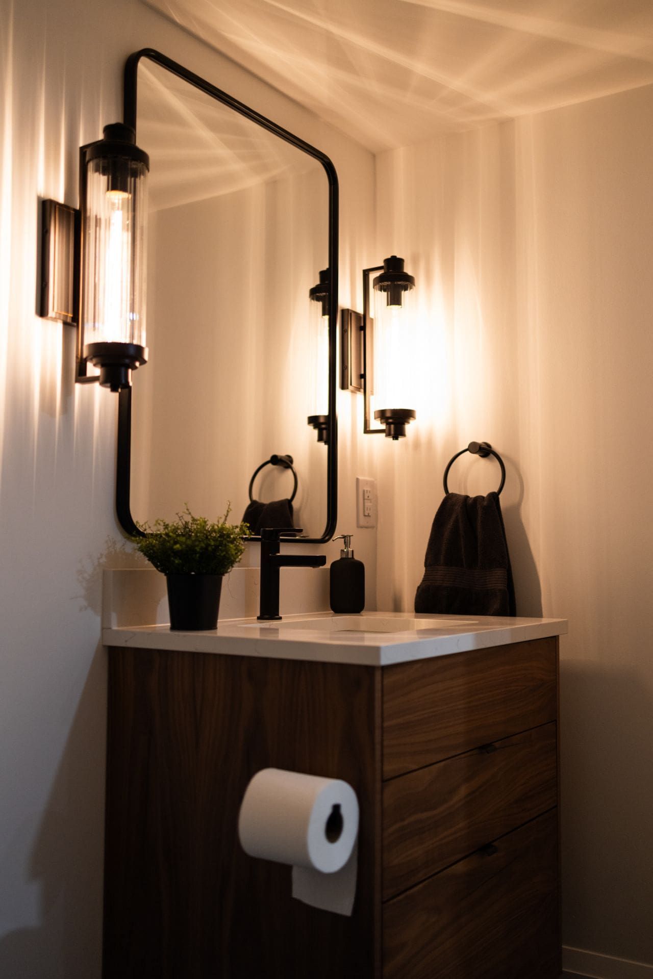 Modern bathroom: wood vanity, mirror with black frame, sconce lighting, and a small potted plant.