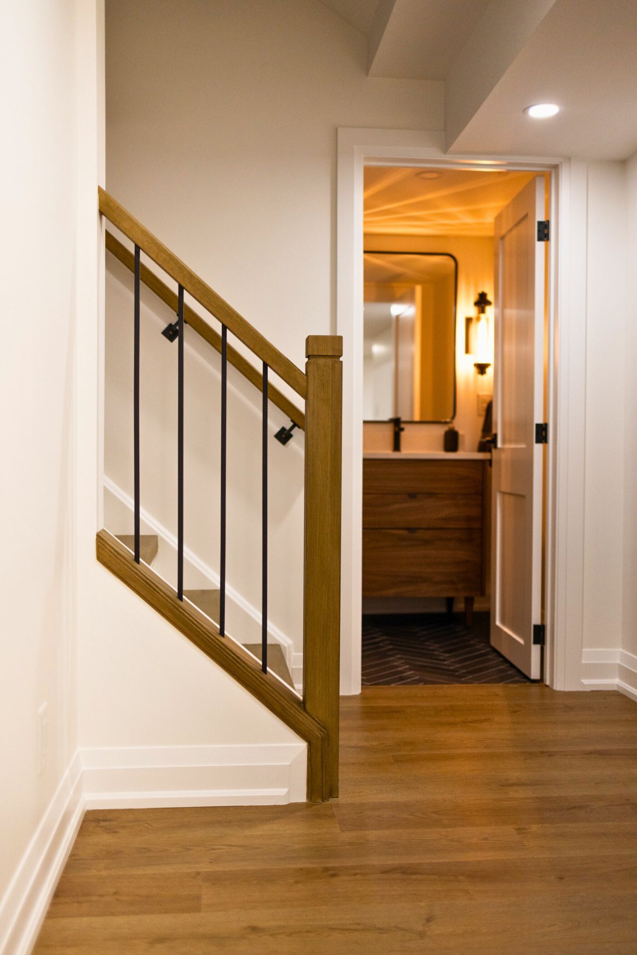 Staircase with wooden handrail, black spindles, leading to a bathroom with wooden vanity and mirror.