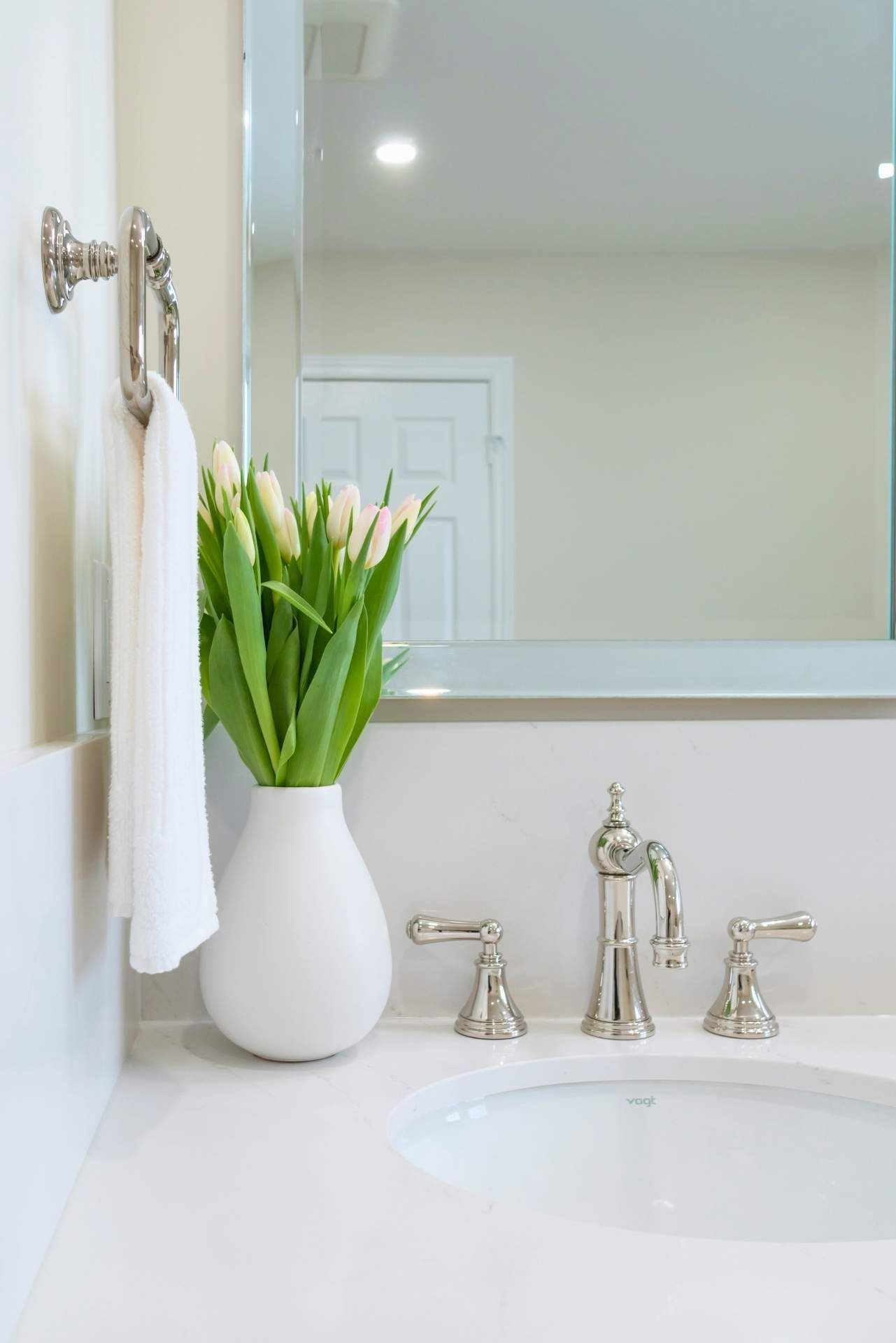 White bathroom with tulips in a vase, towel on a rack, mirror, and faucet.