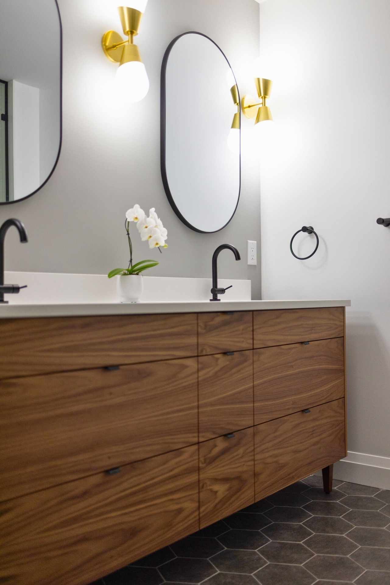 Bathroom with wooden vanity, black fixtures, oval mirrors, and gold sconces; gray walls, orchid.