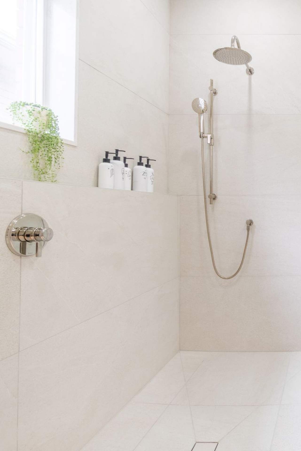 White tiled shower with built-in shelf holding soap bottles, showerhead, and window with plant.