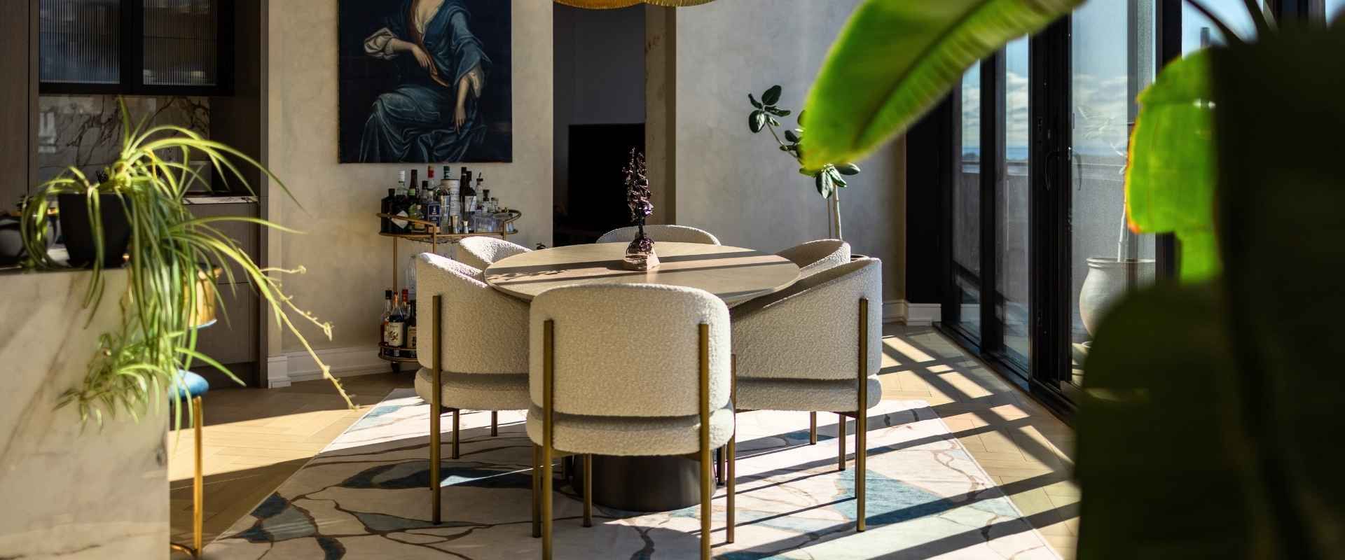 A dining area with a round table, cream upholstered chairs, patterned rug, and a bar cart against a wall with artwork.
