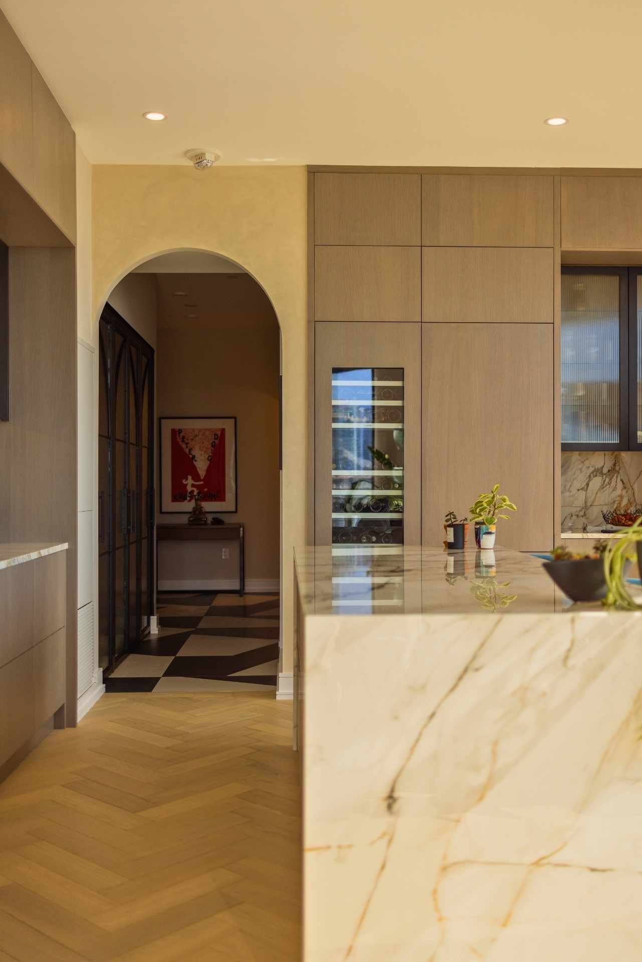 Kitchen with marble island, wine fridge, arched doorway to checkered hallway, and wooden cabinetry.