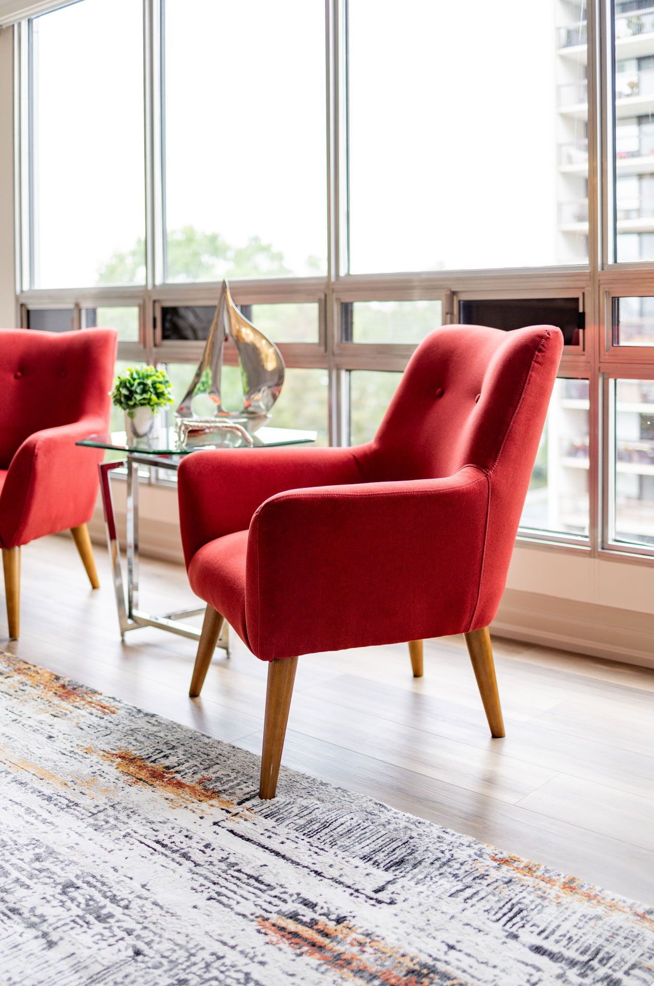 Two red armchairs with wooden legs by a window, near a glass table with a decorative object.