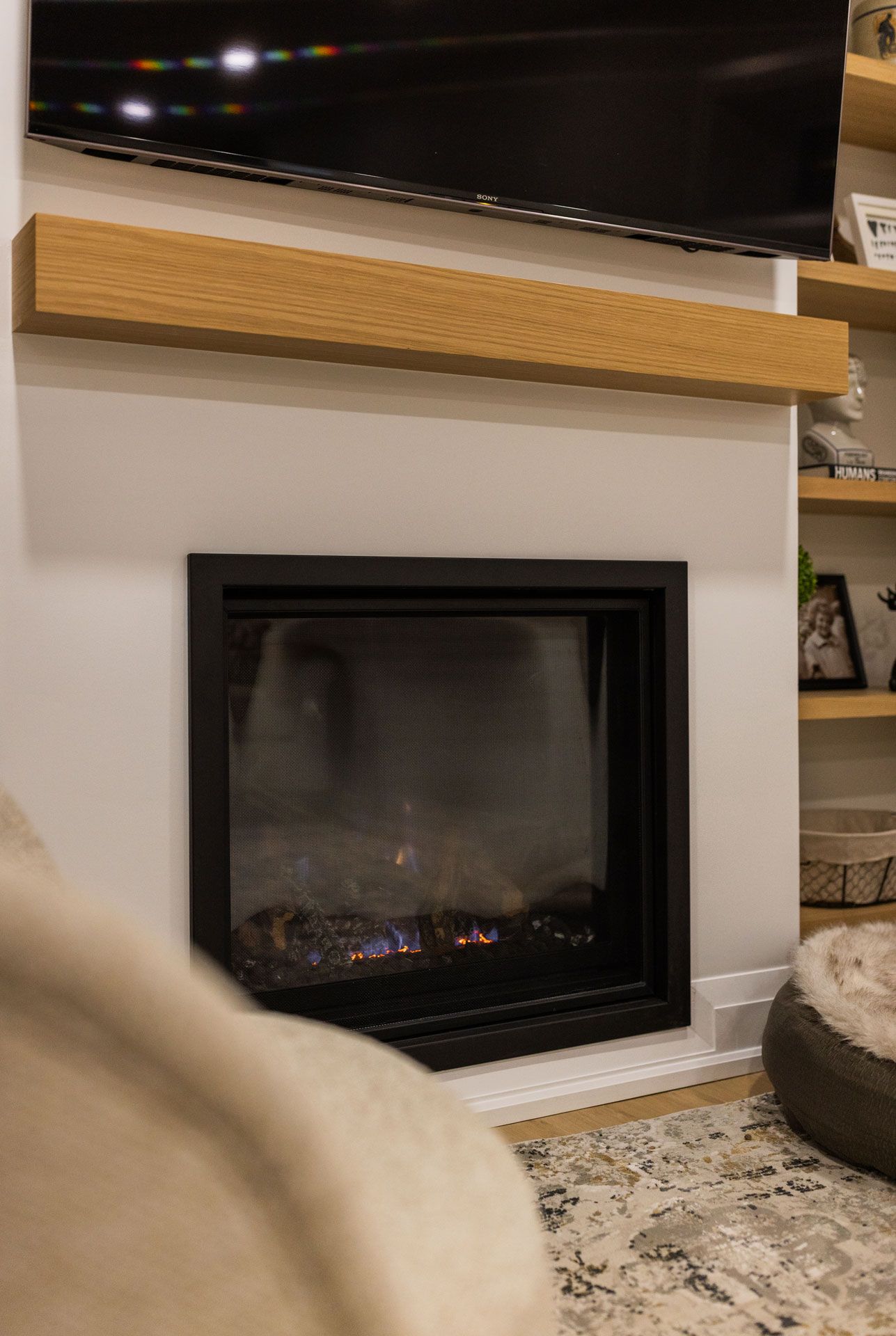 Fireplace with black frame and lit flames, under a wooden mantle and TV, set in a white wall.