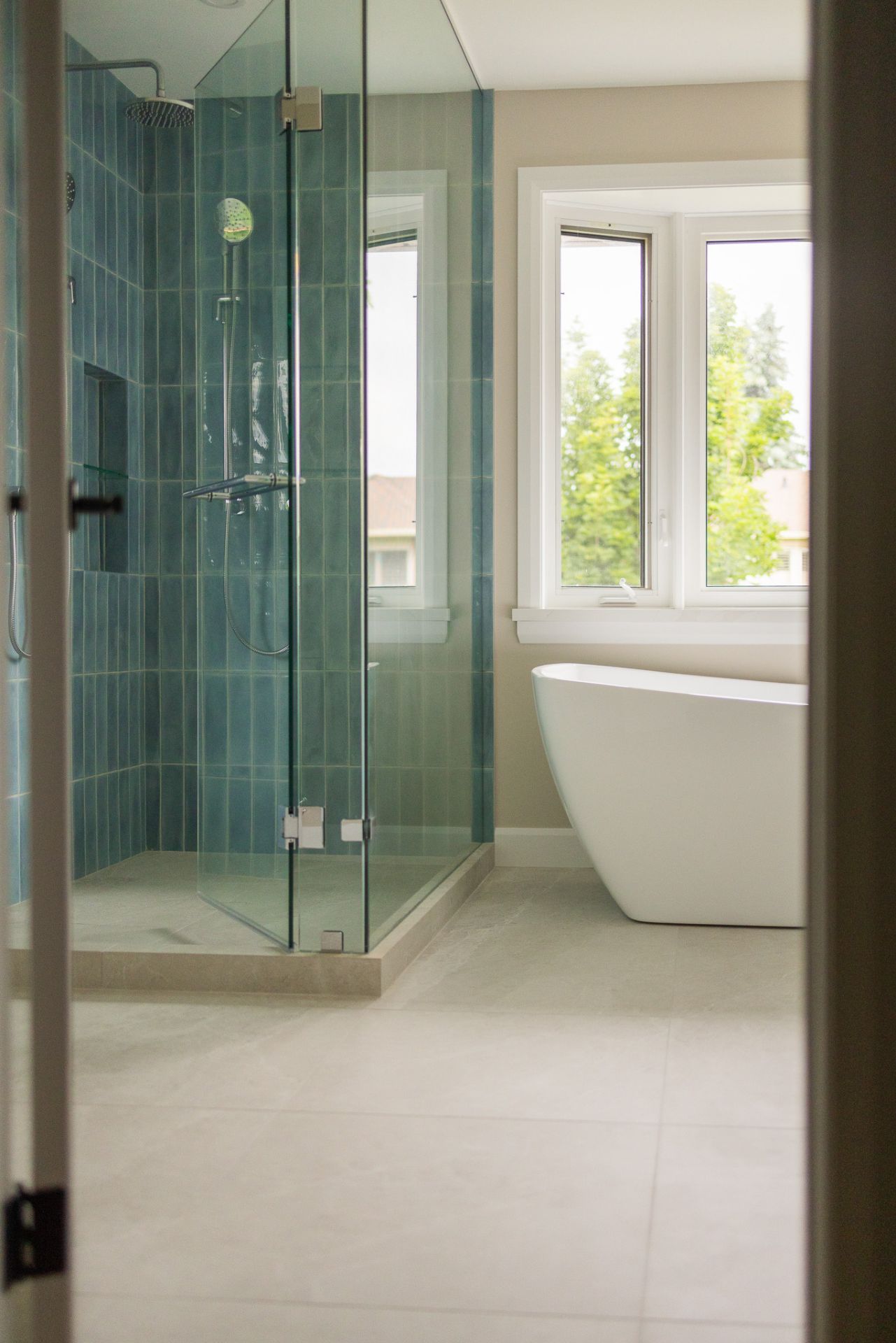 Modern bathroom with teal tiled shower, glass door, white tub, and large window.