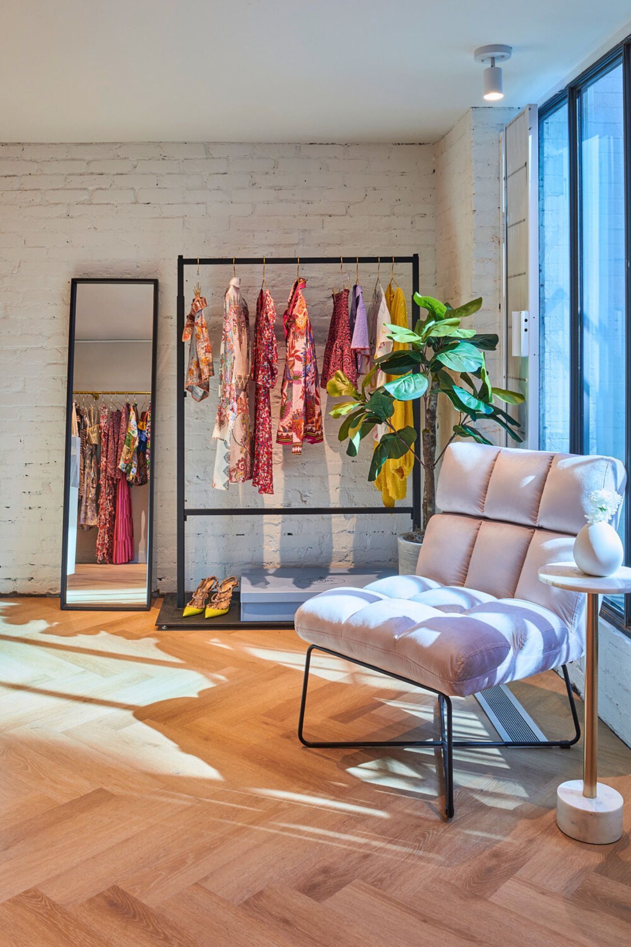 A brightly lit clothing boutique interior with rack of dresses, a chair, mirror, and plant.