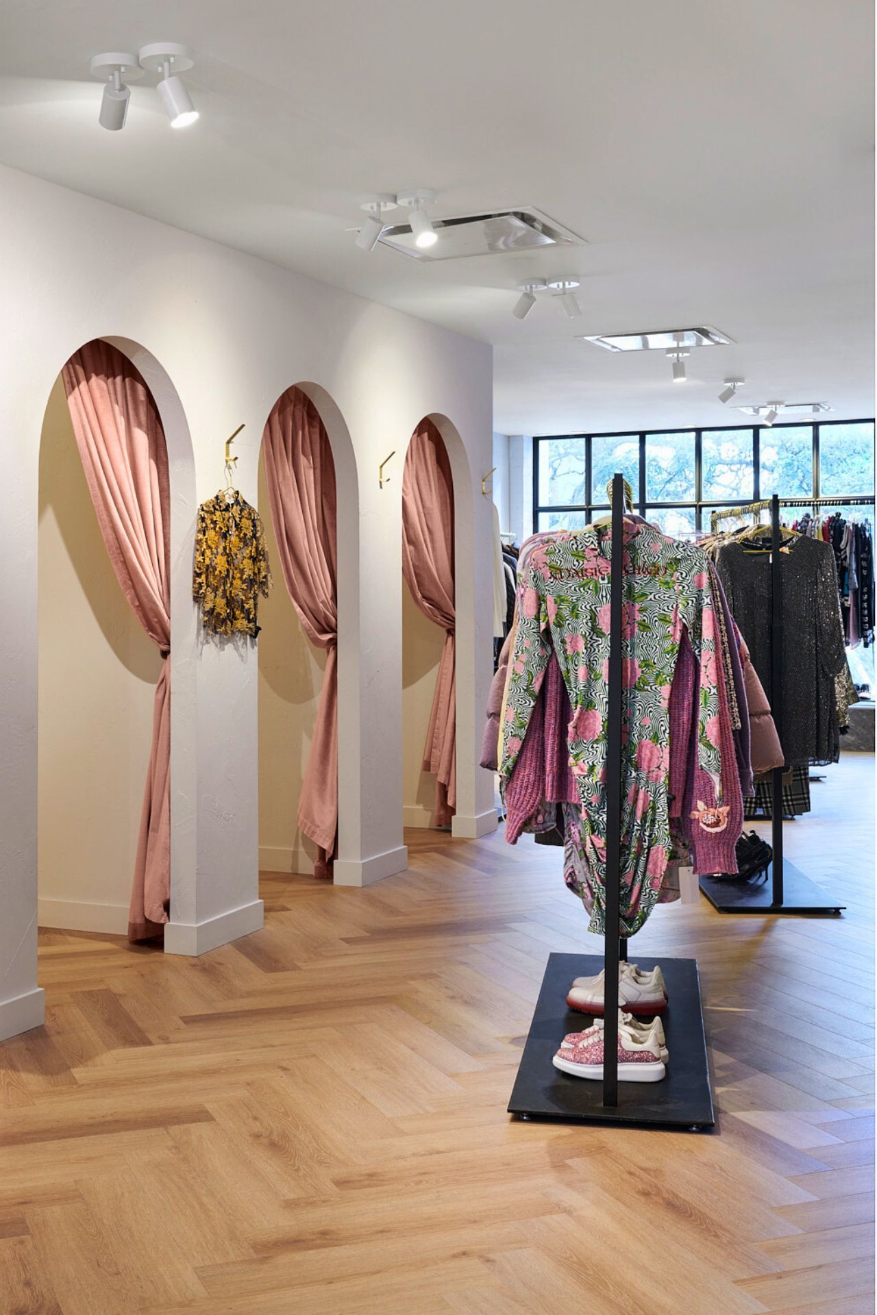 Clothing store interior with arched fitting rooms, pink curtains, and clothes racks. Wooden floor.