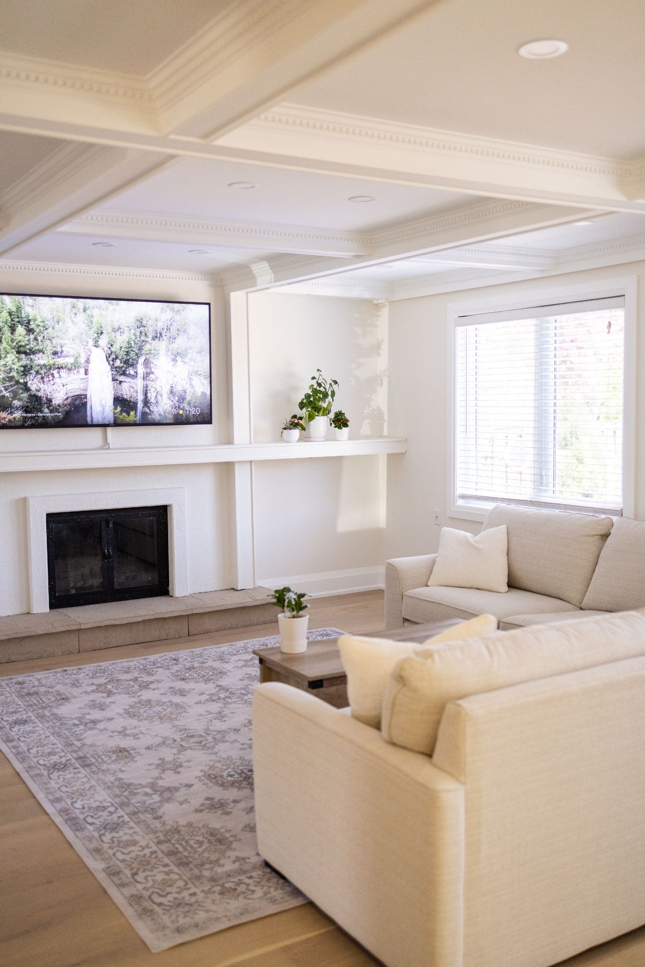 Cozy living room with a white couch, fireplace, TV, and rug. Bright space with wooden floors and a window.