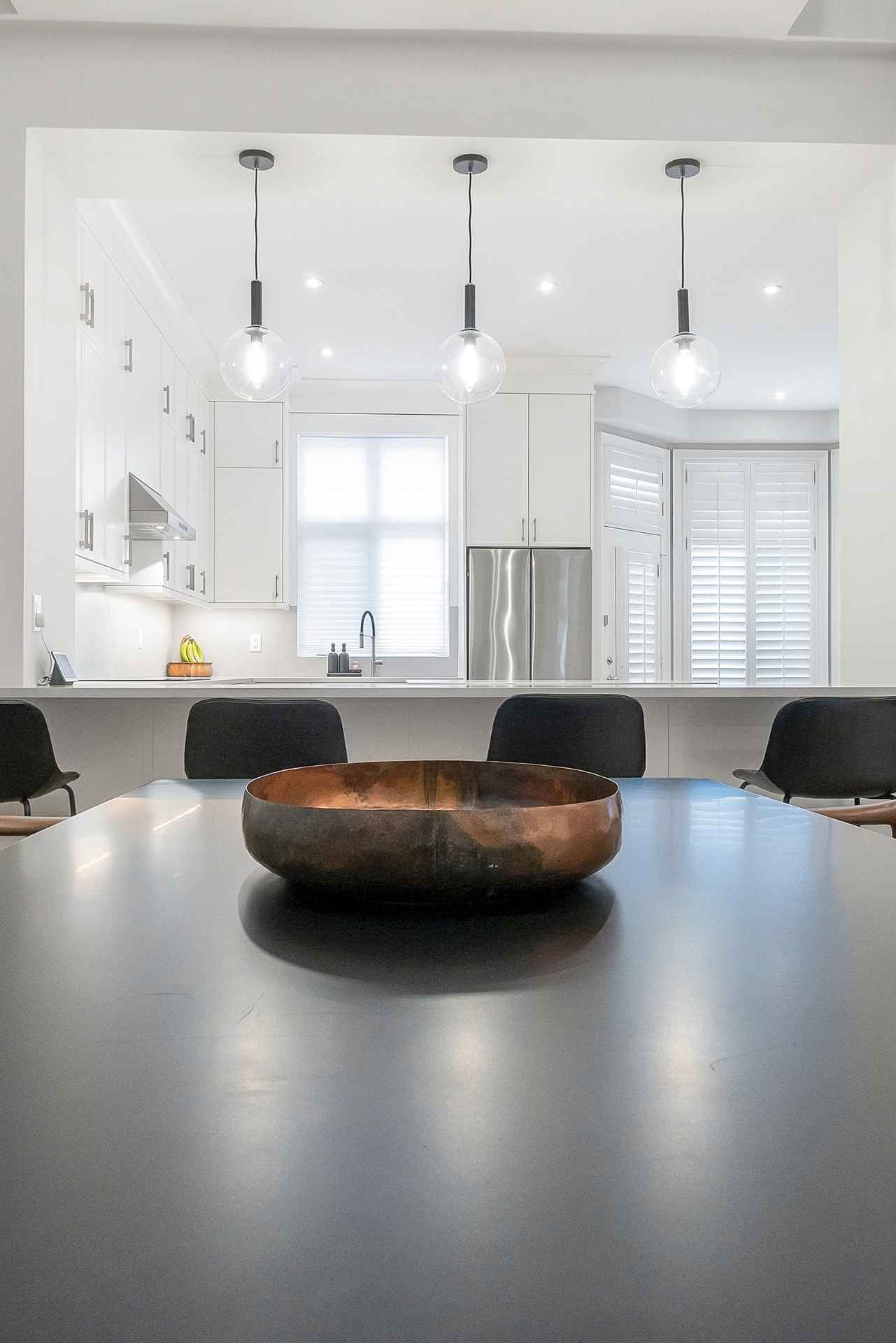 Dining room with dark table, black chairs, and view into a white kitchen with pendant lights.