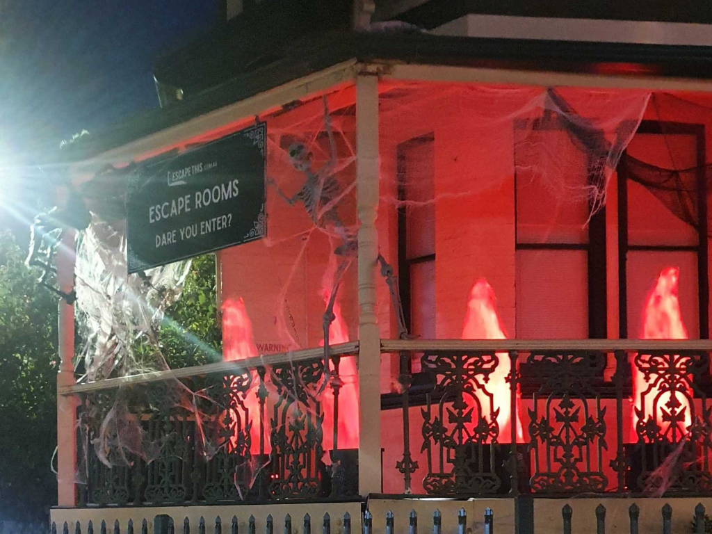Exterior of Escape This Northbridge covered in decorative cobwebs and skeletons for Halloween, lit in red light at night.
