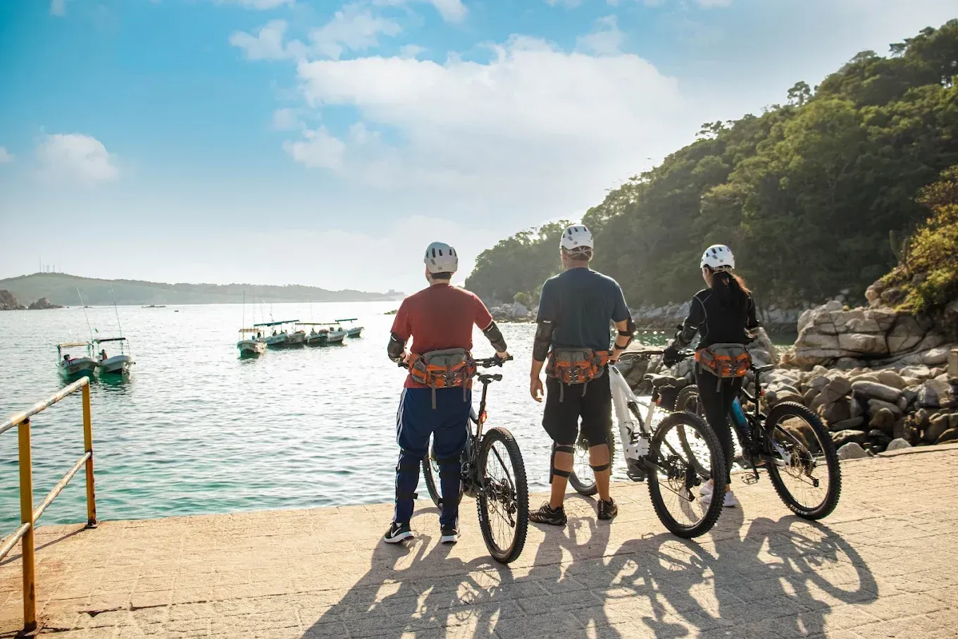 Tres personas en bicicletas, cascos y mochilas están de pie en un muelle, con vistas a una bahía con barcos .