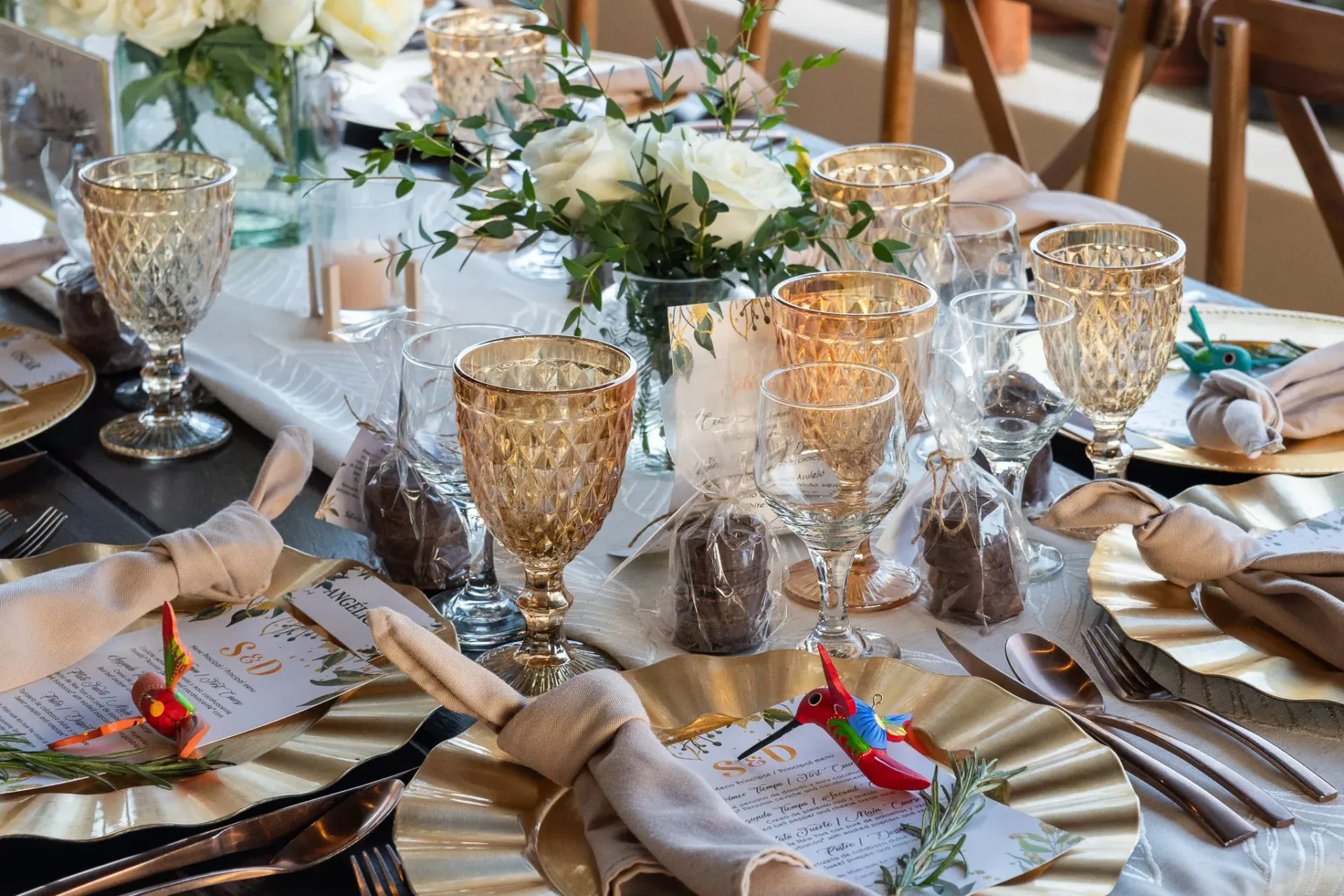 Elegante mesa puesta con platos base dorados, copas ámbar y centros de mesa florales blancos.