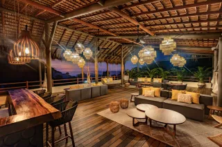 Bar al aire libre con terraza de madera, techo de paja y luces colgantes que se encienden al anochecer. Sofás y mesas