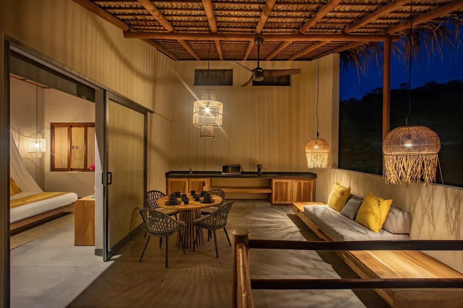 Patio exterior con comedor, asientos y cocina. Muebles de madera, lámparas de mimbre y puerta corredera que da al dormitorio.