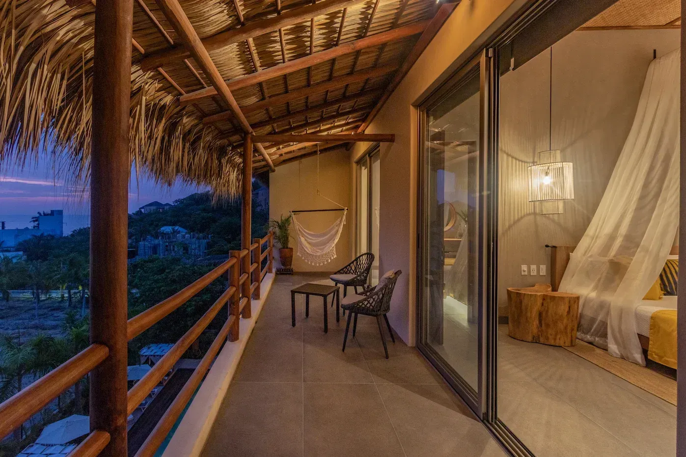 Balcón con techo de paja, con vistas a un paisaje oscuro. Puertas correderas de cristal dan acceso a un dormitorio.