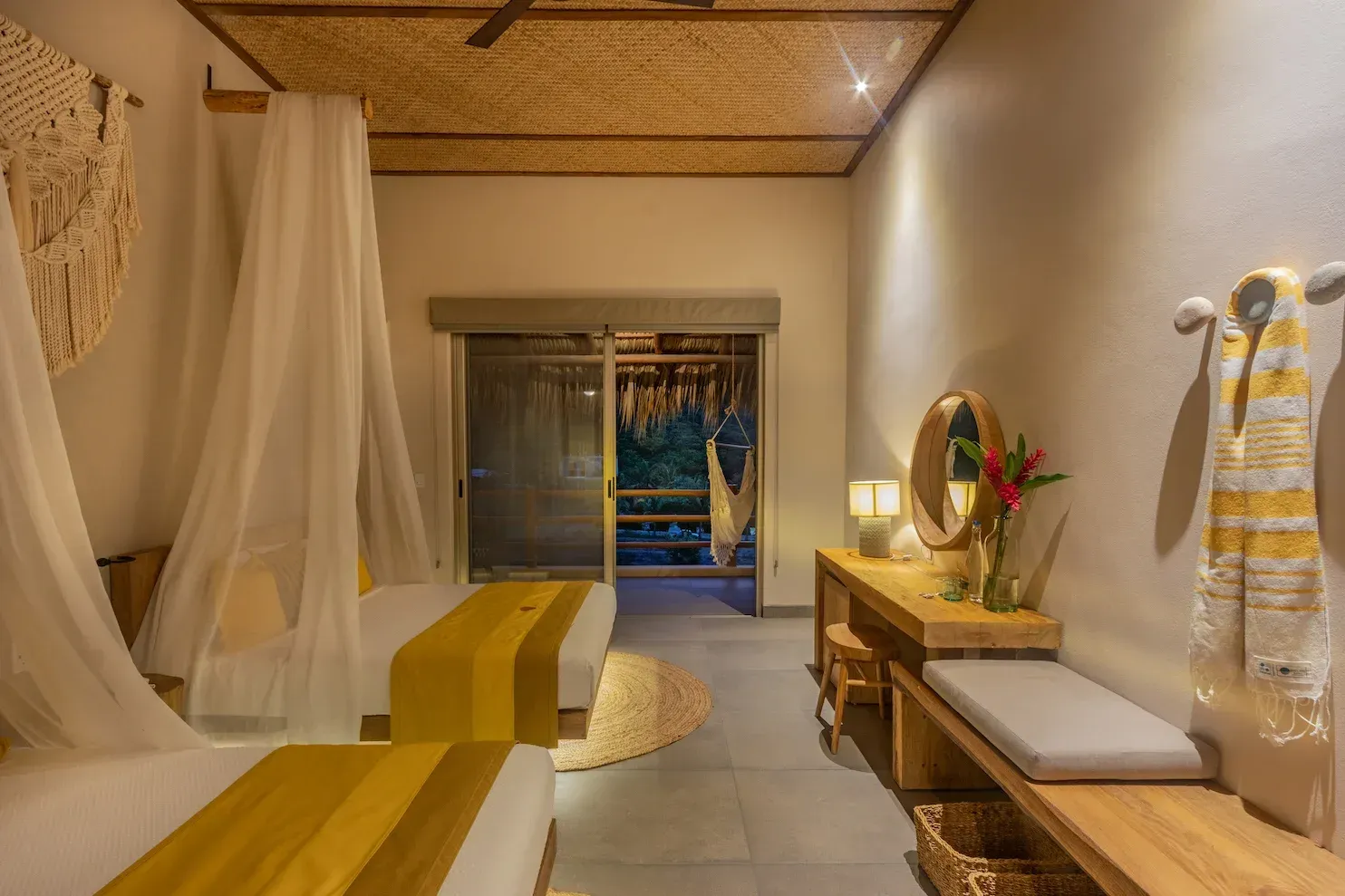 Dormitorio con dos camas, detalles amarillos, cortinas traslúcidas, tocador de madera, puerta abierta al exterior.