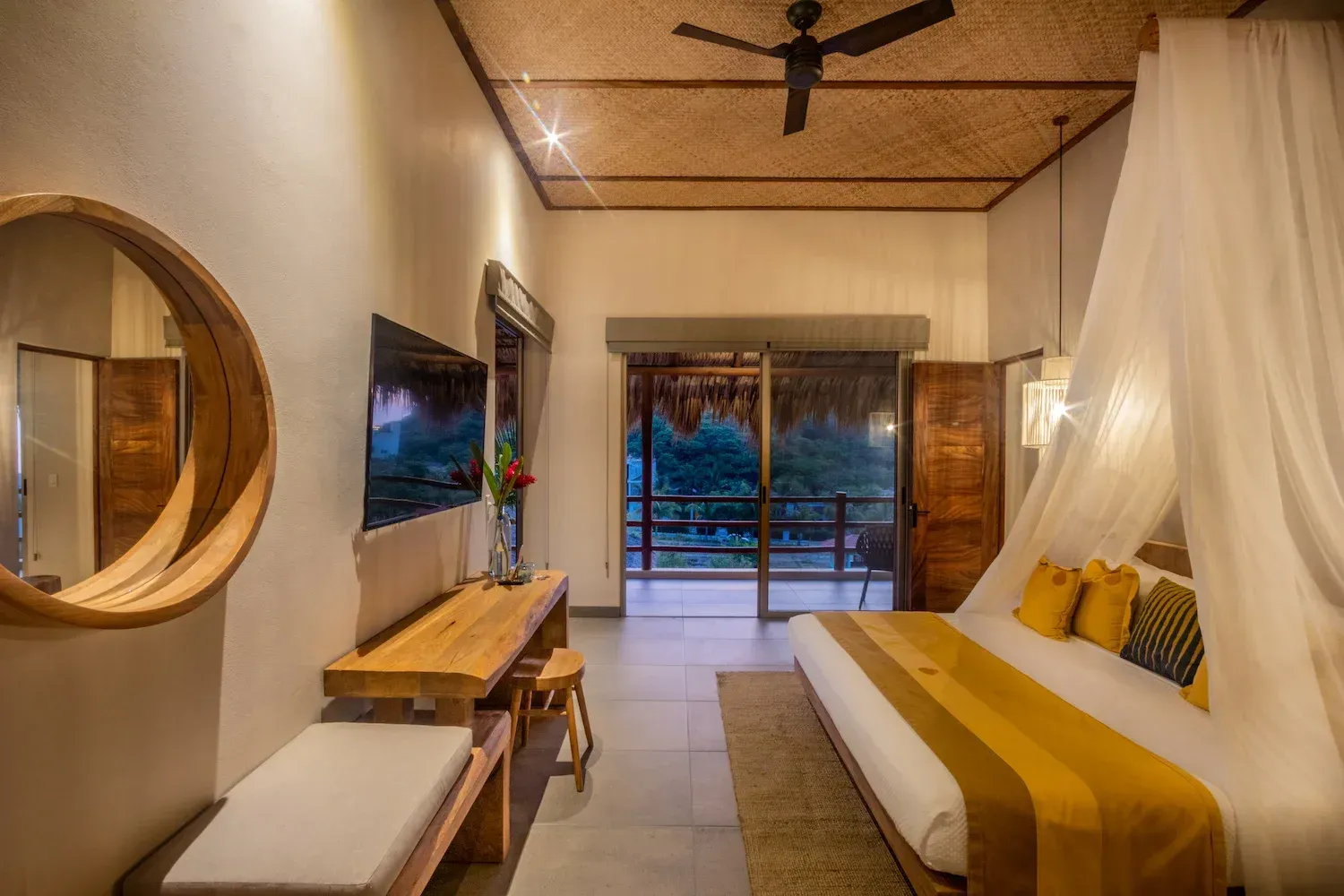 Dormitorio con cama con dosel, escritorio de madera, espejo redondo y balcón con vistas a los árboles.