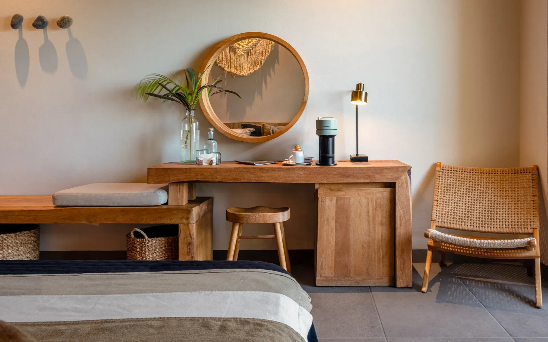 Dormitorio con escritorio de madera, espejo redondo, silla de mimbre y banco.