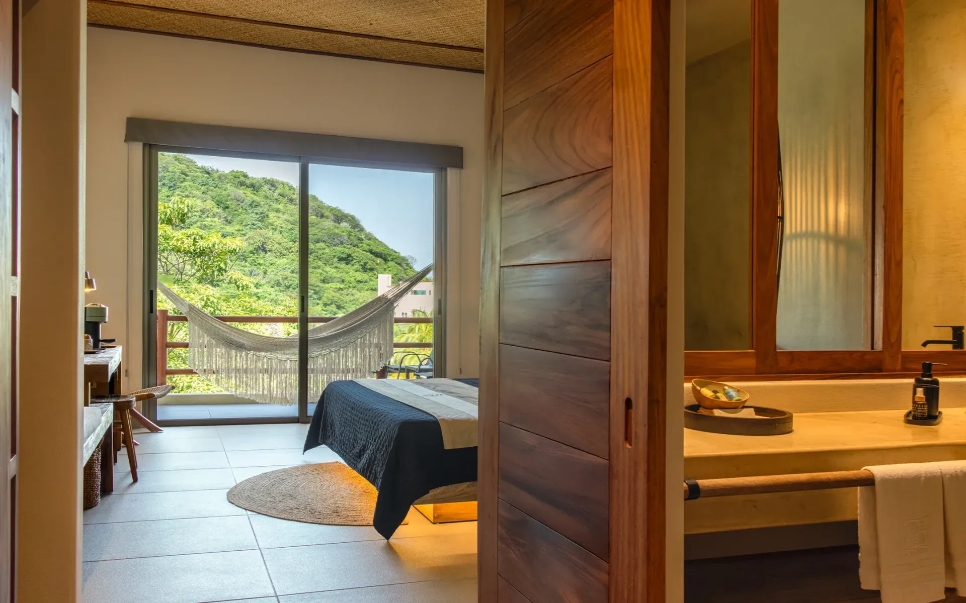 Dormitorio con vistas a la montaña, hamaca en el balcón, puerta de madera abierta al baño, tonos beige y madera.