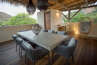 Comedor en la azotea con una mesa larga, sillas y lámparas colgantes. Suelo de madera y mobiliario exterior con vistas.