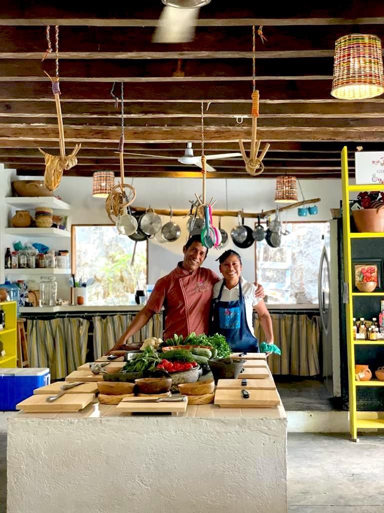 Dos personas de pie en una cocina soleada, sonriendo. Una isla central con para preparar alimentos. Ollas y sartenes .