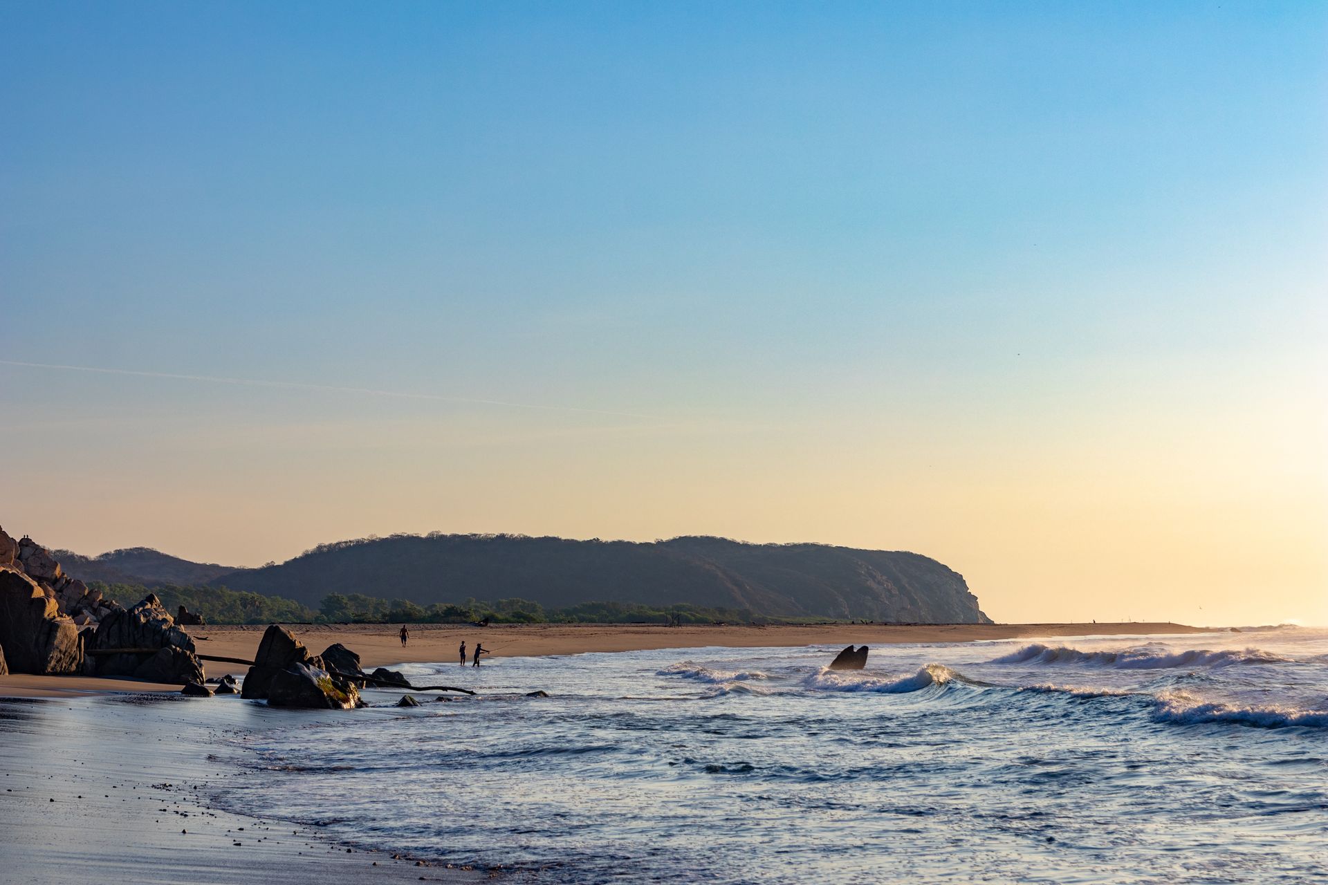 Escena de playa al amanecer: olas, arena, costa rocosa y una colina boscosa a lo lejos bajo un cielo azul pálido y despejado.