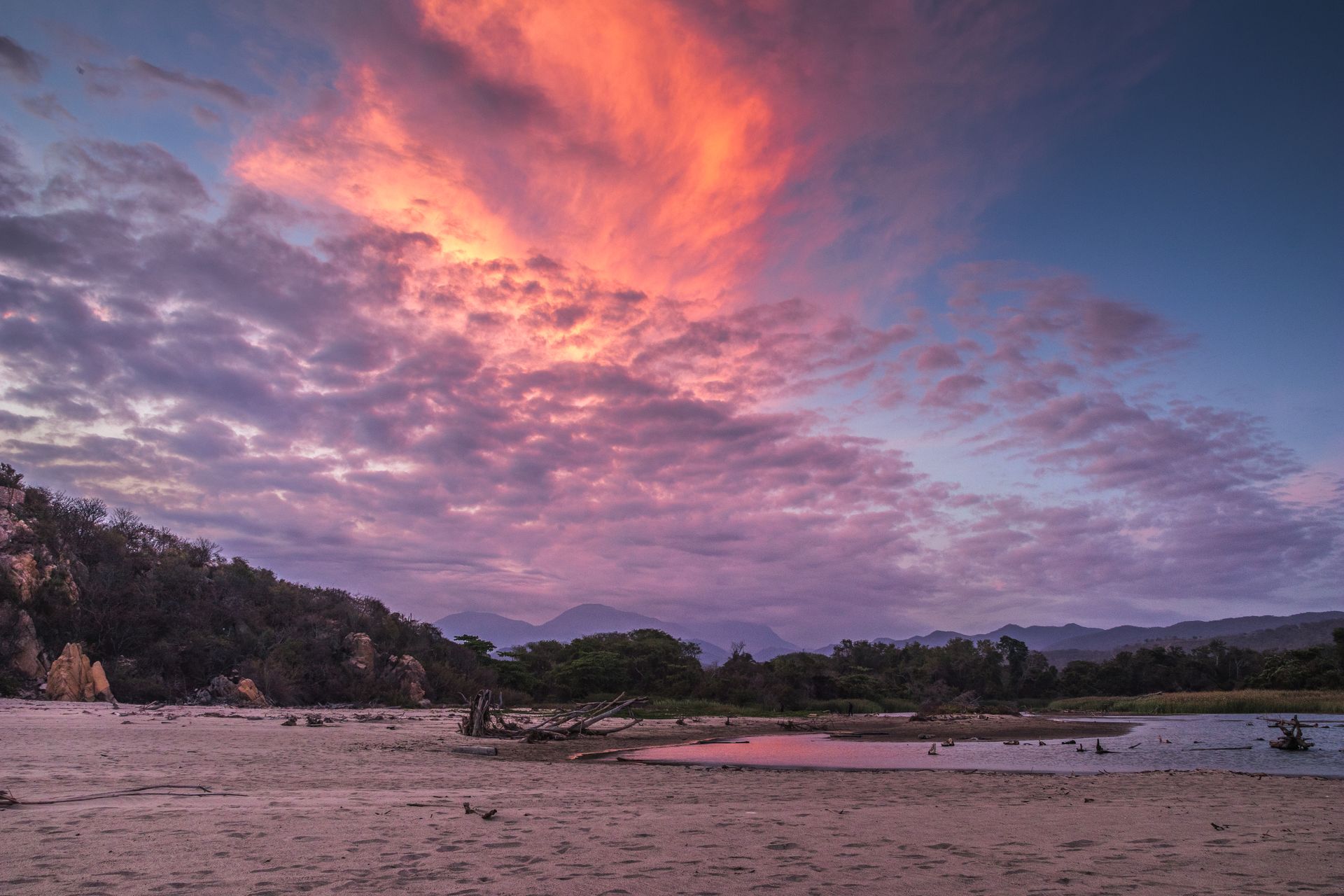 Cielo al atardecer sobre un paisaje con montañas, árboles y una masa de agua poco profunda.