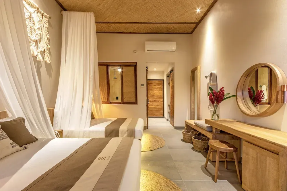 Dormitorio con dos camas, tonos neutros, detalles de madera, cortinas decorativas, espejo redondo y un pequeño tocador.