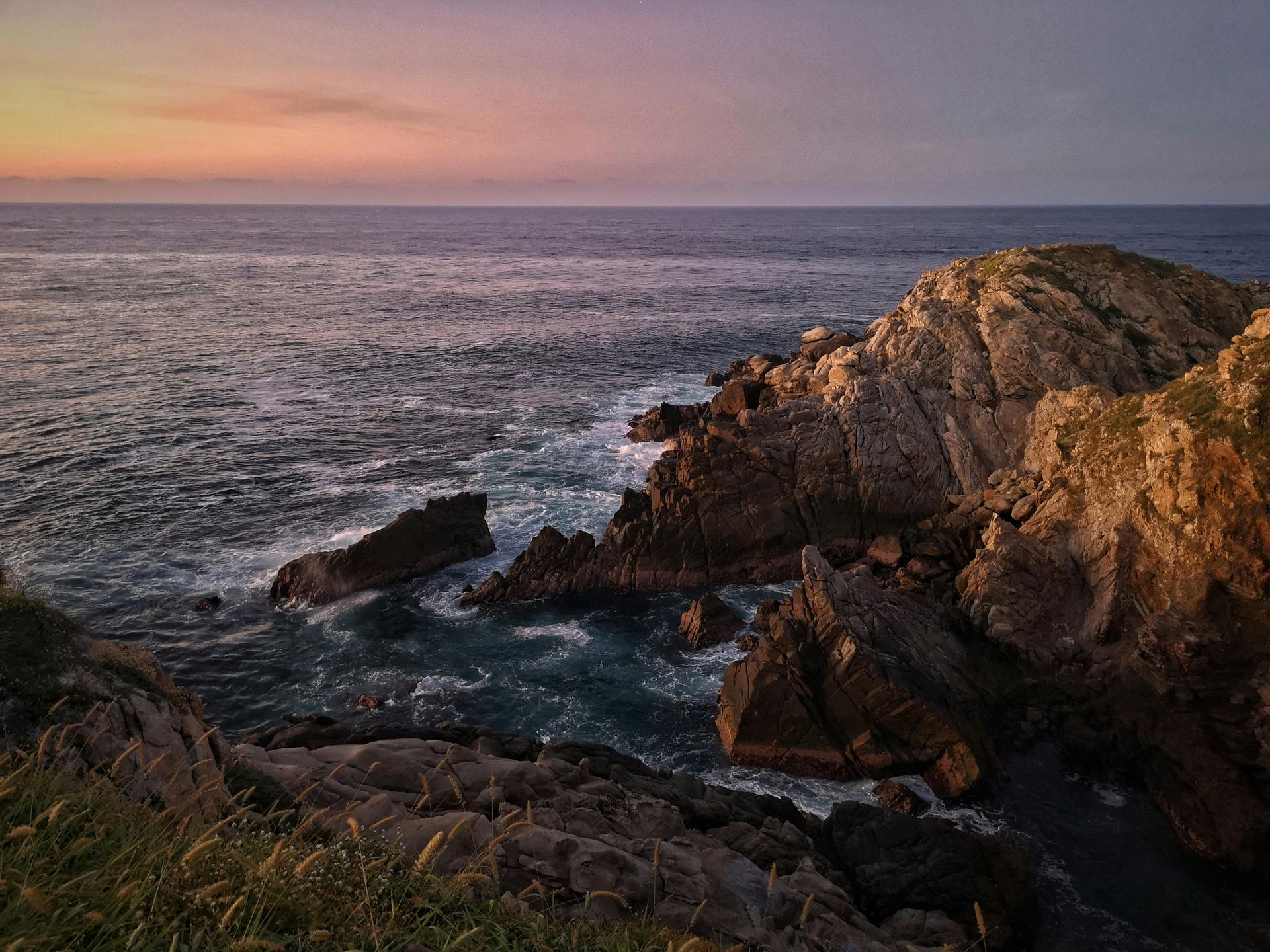 Costa rocosa al atardecer. Las olas del océano rompen contra las rocas oscuras bajo un cielo colorido.